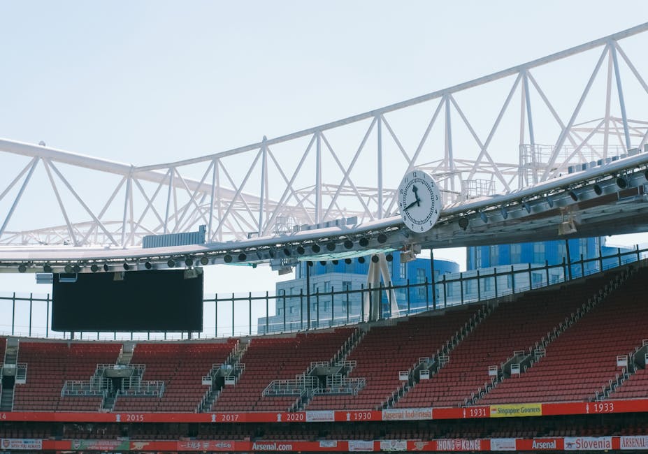 Inside a modern stadium with tiered red seating, a large black electronic scoreboard hangs above the seats, and a round analog clock showing approximately 3:55 is mounted on a white steel structure spanning across the open roof. The structure features a series of crisscross support beams and is visible against a bright sky, indicating clear weather. The stadium area includes a fenced walkway adjacent to the seating section, and the environment appears clean and well-maintained. This scene captures the typical setup of an outdoor sports venue, with the seating area prepared for spectators, and the structural elements designed for viewing and safety. The image could illustrate the setting for a home sports game, event, or sporting fixture.