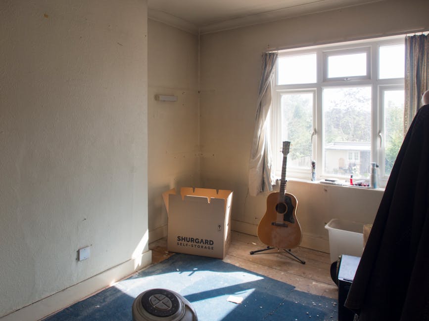 Inside a room designated for home relocation, the image shows a corner near a large window with natural daylight illuminating the space. The cream-colored wall on the left has a mounted electrical outlet near the baseboard. On the wooden floor, there is a blue carpet partially covered by scattered small debris and a portable vacuum cleaner. In front of the window, an empty cardboard box labeled 'SHURGARD SELF-STORAGE' is placed on the floor, indicating packing materials for moving. To the right of the box, a wooden acoustic guitar is positioned upright on a stand, suggesting personal belongings are being packed or prepared for transport. Behind the guitar, there are several small bottles, likely for cleaning or personal care, placed on the windowsill. Adjacent to the windows, a white plastic storage tub is visible, potentially used for packing small items or fragile objects. A dark fabric garment is hanging over a piece of furniture or a coat rack on the far right at the edge of the frame. This scene reflects the early stages of a furniture transport process associated with house removals, with various packing and loading preparations underway, as could be managed by companies like Man with Van Highbury.