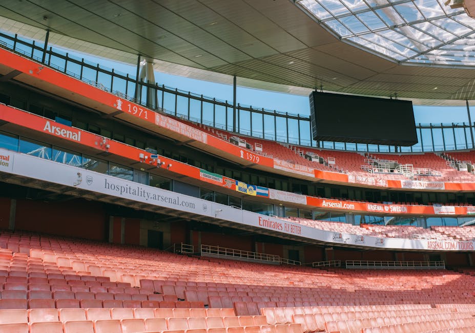 Interior view of the Emirates Stadium in Highbury, showing multiple tiers of red seating arranged in curved rows, with some areas unoccupied. Above the seating, there are advertising banners and a large, empty digital screen hanging from the stadium's roof, which has a modern, curved design with a partially transparent roof covering. In the foreground, the lower tier of seating is visible with a few empty seats, and along the upper tiers, banners display sponsorship and team information. The lighting inside the stadium is natural, with sunlight filtering through the roof panels. This image reflects a sports venue preparing for a match or event, and provides a clear view of the architectural and seating arrangement, relevant for discussions around venue logistics, event planning, or moving and maintenance within large sporting arenas, as managed by companies like Man with Van Highbury.
