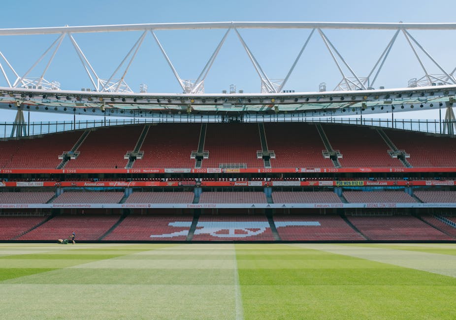 Interior view of the Emirates Stadium in Highbury, showing multiple tiers of red seating arranged in curved rows, with some areas unoccupied. Above the seating, there are advertising banners and a large, empty digital screen hanging from the stadium's roof, which has a modern, curved design with a partially transparent roof covering. In the foreground, the lower tier of seating is visible with a few empty seats, and along the upper tiers, banners display sponsorship and team information. The lighting inside the stadium is natural, with sunlight filtering through the roof panels. This image reflects a sports venue preparing for a match or event, and provides a clear view of the architectural and seating arrangement, relevant for discussions around venue logistics, event planning, or moving and maintenance within large sporting arenas, as managed by companies like Man with Van Highbury.