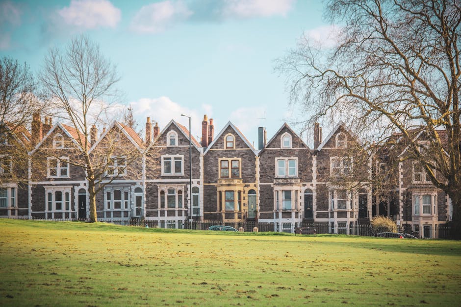A row of Victorian-style terraced houses with stone facades, white window frames, and pitched roofs situated behind a grassy park area. The houses are aligned along a pavement with leafless trees in front, indicating late autumn or winter. The sky is partly cloudy with patches of blue sky visible. This residential scene captures the exterior environment of properties typical in Highbury Fields, relevant to house removals and relocation services. Man with Van Highbury, a professional removals company, might coordinate home relocation or furniture transport within this area, supporting moving logistics and packing processes during house moves. The image displays the outside of the homes, with no moving activity or equipment visible, emphasizing the location context for property handling and transportation in the Highbury N5 postcode.