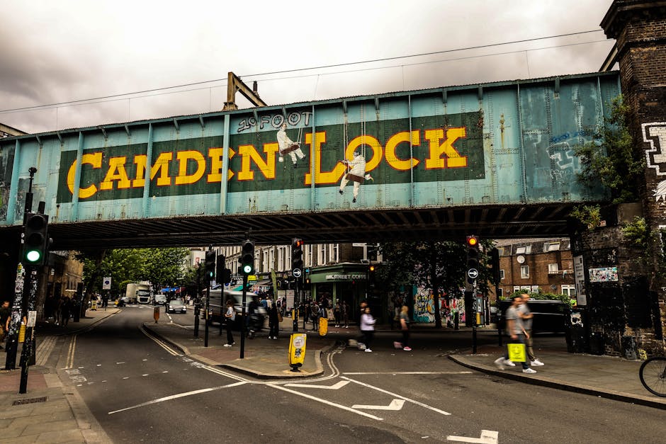 A large weathered railway bridge painted in turquoise, featuring a prominent yellow sign reading 'CAMDEN LOCK' with a painted figure of a girl jumping and holding hands, spans across a busy urban street in Camden Town. Below the bridge, there is a pedestrian crossing with traffic lights, and numerous people walking on the sidewalks and crossing the road. Vehicles, including cars and bicycles, are moving through the intersection, while a collection of shops, graffiti artwork on walls, and trees are visible in the background. The scene captures a typical day of city life with an emphasis on urban infrastructure, outdoor activity, and the vibrant atmosphere of a popular district. This setting is relevant to the context of home relocation or moving logistics as it illustrates a well-known area in North London, supported by signage referencing Camden, which is often associated with local moving and transport activities. Man with Van Highbury may use such imagery to evoke familiarity with neighborhood landmarks involved in the moving process.