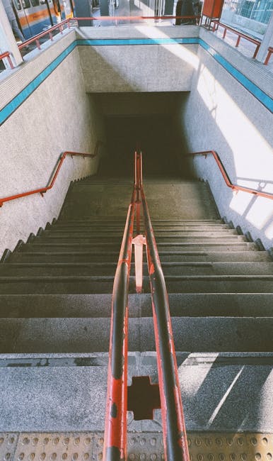 A downward view of a concrete staircase inside a building, featuring brown metal handrails on both sides and a textured, speckled surface on the steps. The staircase leads to an underground level with a rectangular opening at the bottom, revealing a darkened space. The surrounding walls are light-colored with minimal decoration, and natural light filters in from upper windows, partially illuminating the scene. The image depicts an environment suitable for home relocation or furniture transport, with a focus on space and accessibility considerations relevant to various moving logistics, such as narrow staircases that require careful handling during house removals. Man with Van Highbury’s services are relevant for navigating such tight access points during residential moves.