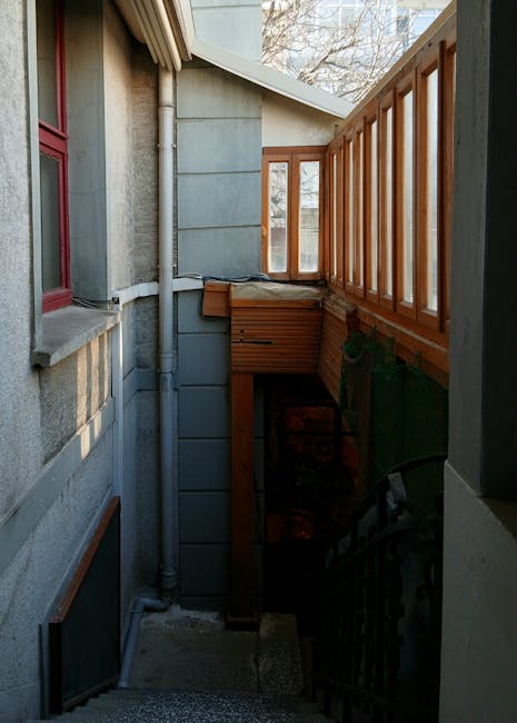 A narrow outdoor staircase leading down from the back of a house, featuring concrete steps with a metal handrail on the right side, adjacent to a wooden balcony with vertical railing panels. The building's exterior wall, visible on the left, is constructed from grey stucco with a red window frame and a small windowsill. A white drainpipe runs vertically along the wall, connecting to the guttering system above. The staircase descends into a small, enclosed space with a wooden storage box positioned at the top of the stairs, partially covered by a protective cloth or padding. Natural daylight illuminates the scene, and the surrounding environment suggests a residential property with trees visible in the background. This image relates to home relocation and furniture transport, illustrating the challenges of moving objects through tight, narrow access points as handled by companies like Man with Van Highbury during packing and loading processes.