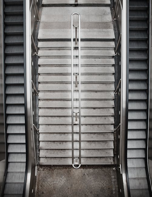 A downward view of a concrete staircase inside a building, featuring brown metal handrails on both sides and a textured, speckled surface on the steps. The staircase leads to an underground level with a rectangular opening at the bottom, revealing a darkened space. The surrounding walls are light-colored with minimal decoration, and natural light filters in from upper windows, partially illuminating the scene. The image depicts an environment suitable for home relocation or furniture transport, with a focus on space and accessibility considerations relevant to various moving logistics, such as narrow staircases that require careful handling during house removals. Man with Van Highbury’s services are relevant for navigating such tight access points during residential moves.