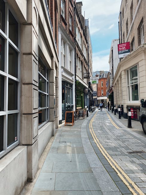 A narrow commercial street with a curved pavement lined by multi-storey buildings on both sides. On the left, there are large windows with metal frames, and on the right, there are smaller windows with white frames and a white facade. Several 'to let' signs are visible hanging above the street, indicating available retail spaces. The pavement is made of grey and light-colored stone tiles, with a double yellow line running along the curb, suggesting parking restrictions. A few pedestrians are walking further down the street, some near small outdoor seating arrangements outside a green shop. The sky is partly cloudy, casting natural light on the scene. During a house or commercial relocation, Man with Van Highbury might be involved in transporting furniture or boxes through streets like this as part of their removal services, with careful navigation along the sidewalk and street features visible in the photograph.