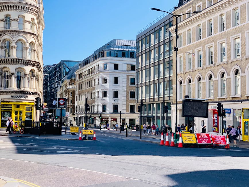 A busy urban street scene in Highbury with a mix of modern and historic buildings lining the pavement, featuring a narrow road temporarily closed for access with red and white traffic cones, barriers, and road signs indicating restrictions for through traffic and access only. Pedestrians are walking along the sidewalk, some waiting near the entrance of a building, while others cross the street. On the left side, there is a storefront with a yellow sign and a person walking past. In the foreground, a section of the road shows markings for vehicle control, and the area is illuminated by a streetlamp. This setting illustrates a typical location where house removals and furniture transport activities would take place, with [COMPANY_NAME] potentially involved in managing the logistics of loading or unloading goods during a home relocation or moving project, aligned with the services in the specified [PAGE_TITLE].
