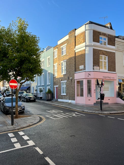 A narrow commercial street with a curved pavement lined by multi-storey buildings on both sides. On the left, there are large windows with metal frames, and on the right, there are smaller windows with white frames and a white facade. Several 'to let' signs are visible hanging above the street, indicating available retail spaces. The pavement is made of grey and light-colored stone tiles, with a double yellow line running along the curb, suggesting parking restrictions. A few pedestrians are walking further down the street, some near small outdoor seating arrangements outside a green shop. The sky is partly cloudy, casting natural light on the scene. During a house or commercial relocation, Man with Van Highbury might be involved in transporting furniture or boxes through streets like this as part of their removal services, with careful navigation along the sidewalk and street features visible in the photograph.